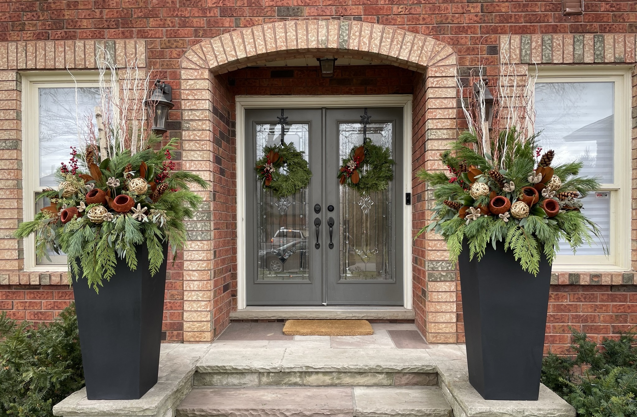 Arched brick entry with matching evergreen wreaths and two tall winter planters of cedar, birch and red accents
