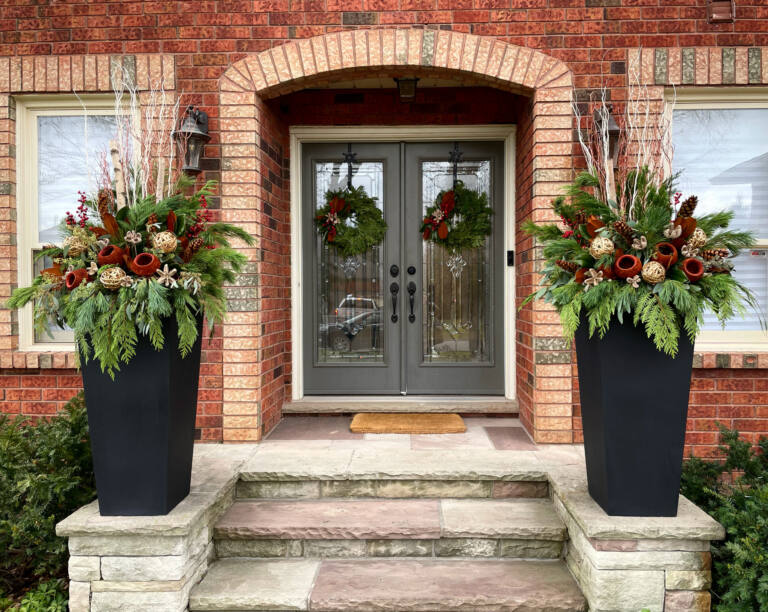 Two tall black planters with red and green winter arrangements at a brick arched double door