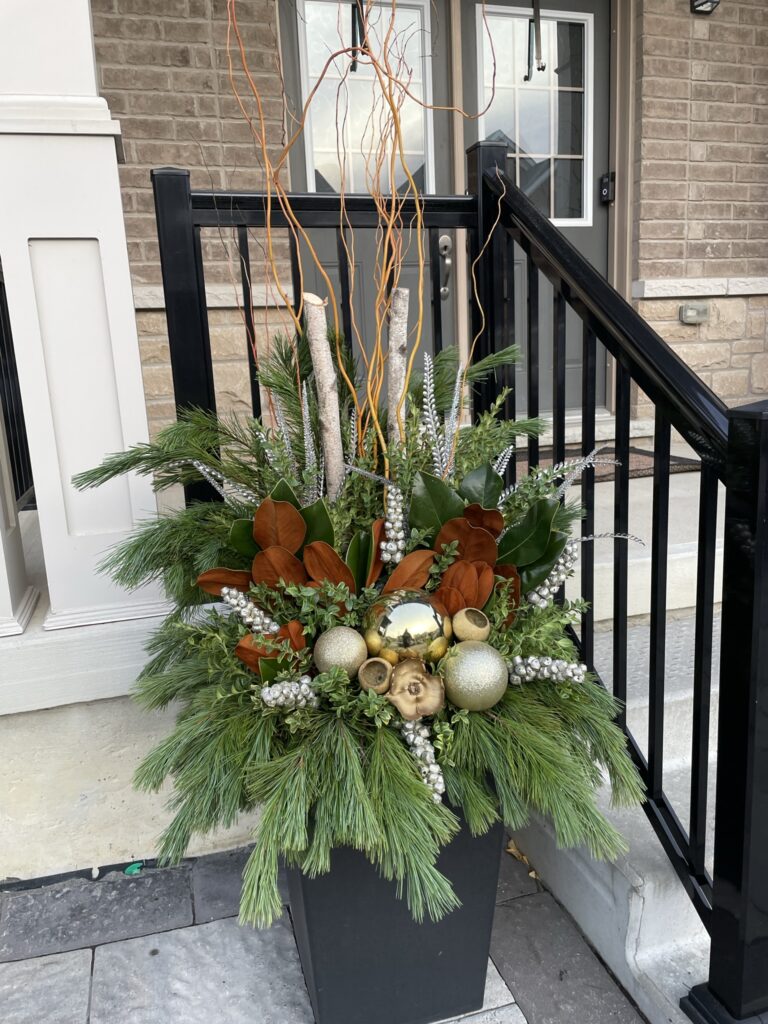 Winter planter with white birch poles, gold ornaments and cascading cedar beside a black railing