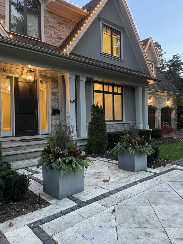 Modern grey rectangular planters flanking the covered entry of a stone and shingle house at dusk