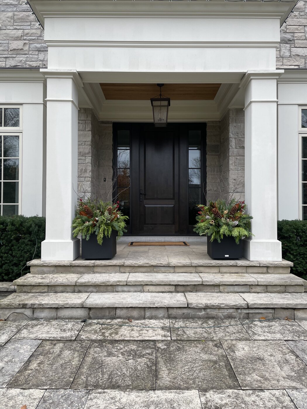 A square black planter filled with cedar, pinecones and birch beside a white column