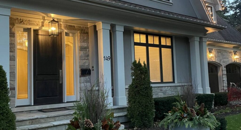 A decorated front entrance at dusk with lit windows and two winter planters
