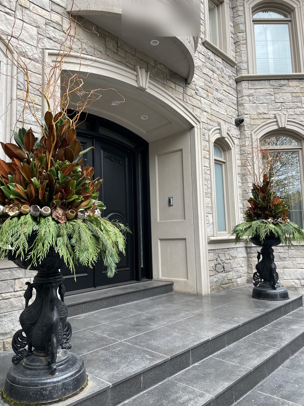 Tall black urns with magnolia and cedar framing the arched entrance of a stone home